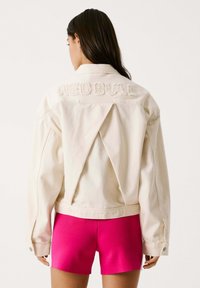 Chaqueta de denim crema con un corte distintivo en la parte posterior. Presenta el texto bordado "RED BULL" en la parte posterior y mangas largas. Combinada con pantalones cortos rosas.