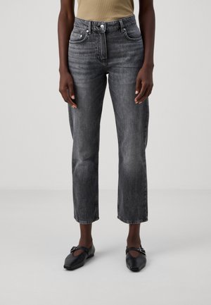 Personne portant un jean gris taille haute à jambes droites, un haut beige et des ballerines noires pointues avec des boucles, debout devant un fond clair uni.