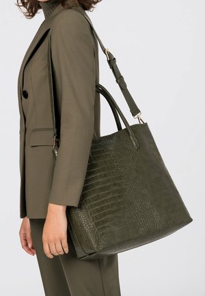 Mujer con blazer color oliva llevando un gran bolso tote de cuero verde oscuro con estampado de cocodrilo y asas sobre el hombro.