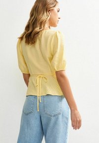 Gul, texturerad blus med puffärmar och knytbakdetalj, tillsammans med ljusblå högmidjade jeans. Bakvy visas.