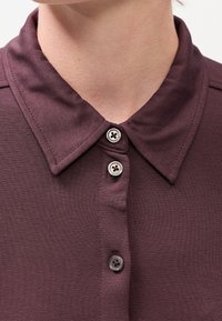 Gros plan d'une personne portant une chemise col montant de couleur violet foncé avec trois boutons argentés visibles boutonnés.