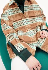 Person som bär en brun och vit rutig skjorta över en mörkgrön långärmad tröja, sittande på en grön stol med händerna vilande i knät.