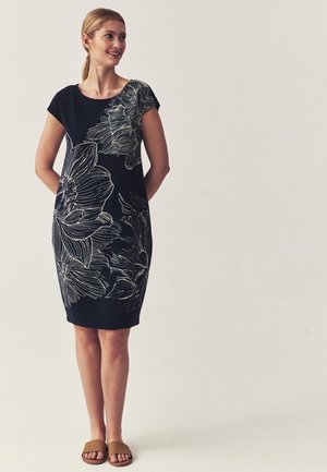 Kobieta stoi z rękami za plecami, ubrana w czarną sukienkę z dużym białym kwiatowym wzorem i brązowe klapki typu slide.