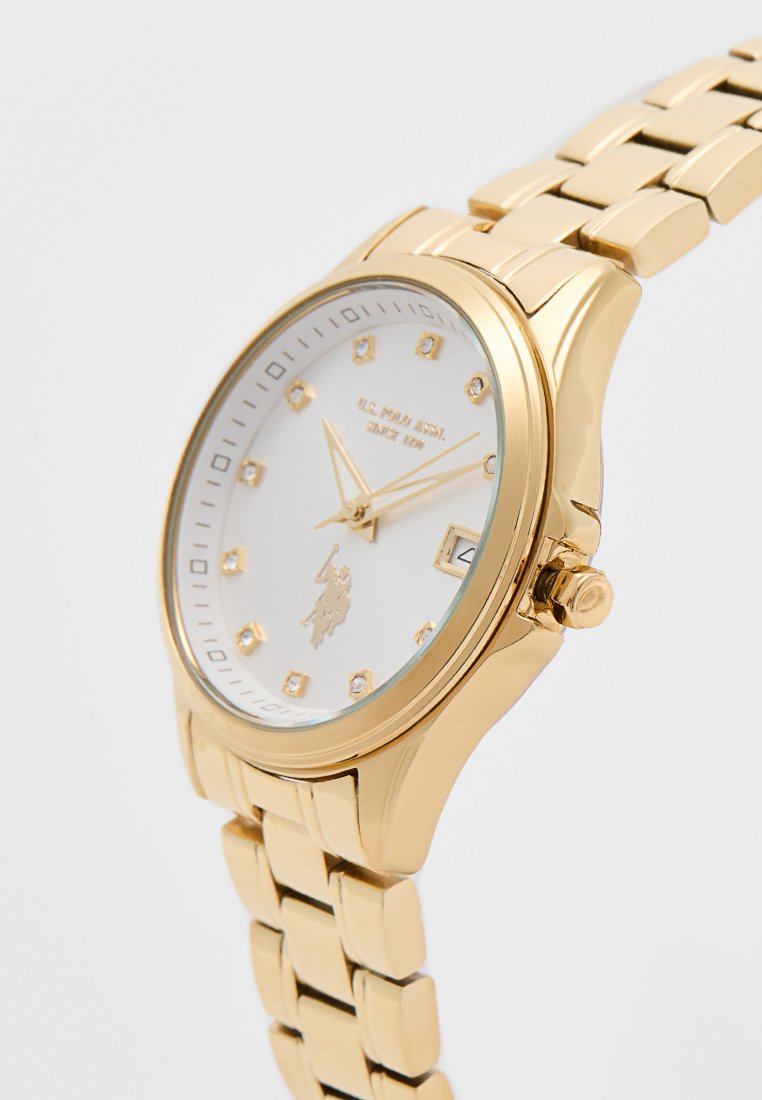Reloj de pulsera de metal dorado con esfera blanca, marcadores de hora de cristal, pantalla de fecha y logotipo y texto de U.S. Polo Assn. en la cara.