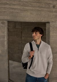 Jasnoszary sweter z zamkiem pod szyją i ściągaczami na rękawach, zestawiony z turkusową koszulą i niebieskimi dżinsami. Model trzyma czarny plecak. Tło betonowe.