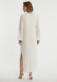Lange witte jurk met ingewikkelde diamanten patronen, lange mouwen en zijsplitten, gemaakt van een lichte, gestructureerde stof.