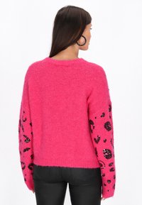 Puszysty różowy sweter z czarnym cętkowanym wzorem w panterkę na rękawach, prążkowany okrągły dekolt i luźny krój, zestawiony z czarnymi spodniami.