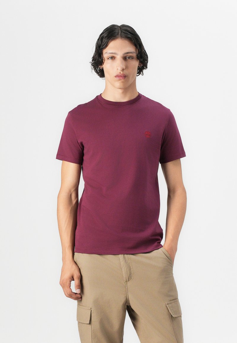 T-shirt in cotone bordeaux con collo rotondo. Presenta un piccolo logo rosso sul lato sinistro del petto. Il modello indossa pantaloni khaki e sta in piedi davanti a uno sfondo bianco.