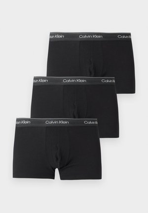 Trois boxers noirs Calvin Klein avec des ceintures grises présentant des logos blancs Calvin Klein, disposés en chevauchement sur un fond blanc.