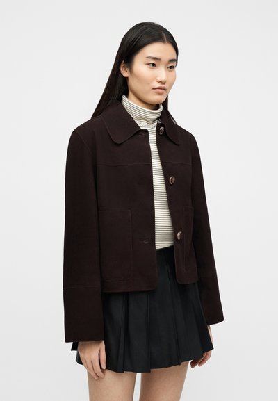 Γυναίκα που φοράει σκούρο καφέ σακάκι με κουμπιά, μαύρη πλισέ φούστα και λευκό ζιβάγκο με ρίγες, στέκεται μπροστά σε απλό ανοιχτό φόντο.