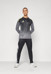 Athletische outfit met een donkergrijze lange mouw top met lichtgrijze en neongele accenten, gecombineerd met zwarte broek met witte zijstrepen.