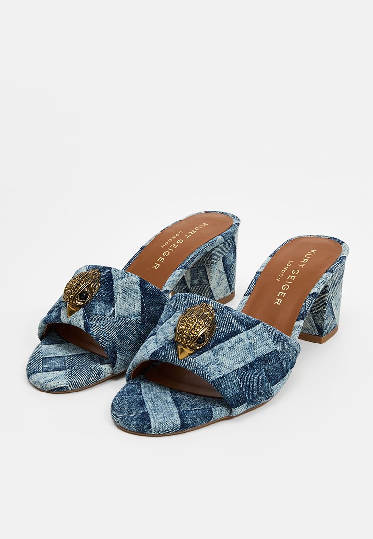 Mules de mezclilla azul con una textura trenzada, diseño de punta abierta y un acento decorativo dorado en la parte superior. Tacón cuadrado y grueso.