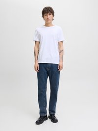 Bijela majica s okruglim izrezom u kombinaciji s plavim trapericama. Model nosi crne cipele s vezicama, stojeći na neutralnoj pozadini.