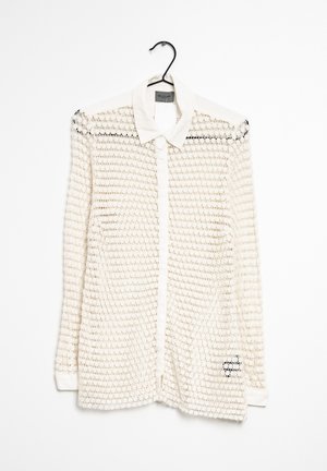 Valkoinen pitkähihainen nappikauluspaita, jossa on terävä kaulus ja virkattu avoin kudoskuvio, roikkuu mustassa hengarissa valkoista taustaa vasten.