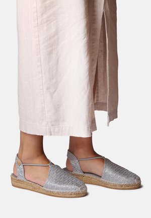 Feet wearing silver woven espadrille sandals with ankle straps, paired with light beige wide-leg cropped pants with side slits.