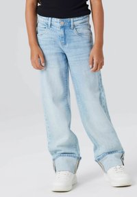 Barn som har på seg lyseblå vide jeans med oppbrettede buksebein og hvite joggesko, stående mot en nøytral bakgrunn.