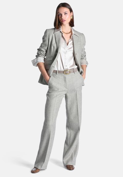 Femme debout les mains dans les poches, portant un costume texturé gris clair, une blouse blanche en satin, une ceinture beige, un collier en or, des boucles d'oreilles et des chaussures à motifs.