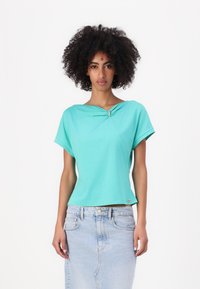 T-shirt turchese a maniche corte con scollatura arricciata e dettaglio metallico decorativo. Tessuto in cotone morbido abbinato a una gonna di jeans azzurri.