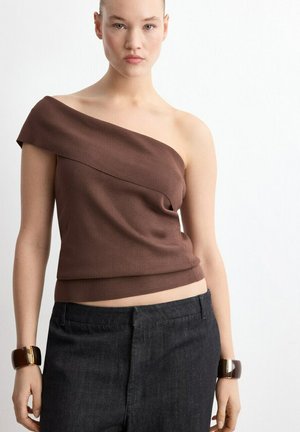 Ung kvinde iført en brun top med én skulder og mørke bukser, stående foran en ensfarvet hvid baggrund med armbånd på begge håndled.