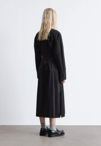 Schwarzes, langärmliges Kleid mit eng anliegendem Taillenbereich, lockerem Rock und offenem Rückendetail. Kombiniert mit schwarzen Schuhen und grauen Socken. Minimalistisches Design.