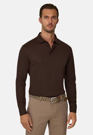 Hombre con camisa tipo polo de manga larga color marrón oscuro, pantalones beige y un cinturón marrón trenzado sobre fondo liso.