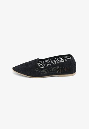 Zapato plano de encaje negro con punta puntiaguda y suela beige, con patrones florales en el diseño del encaje.