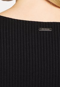 Sort ribbet top med en bred halsudskæring og et metallic "GUESS" mærke bagpå. Teksturen er blød, og stoffet ser ud til at være strækbart.