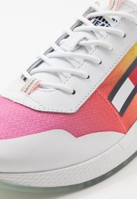 Gros plan d'une basket blanche avec un bout texturé rose, des lacets blancs et un motif à rayures multicolores sur le côté.