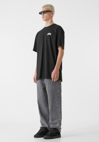 Melna pārmērīga T-kreklu ar baltu logo, saskaņots ar pelēkiem džinsiem un melniem sporta apaviem. Modelis valkā tumšas taisnstūrveida saulesbrilles.