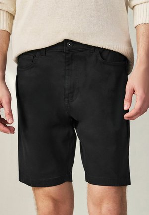 Muž v čiernych šortkách s dĺžkou po kolená a bočnými vreckami, oblečený v béžovom pletenom svetri, stojaci na neutrálnom pozadí.