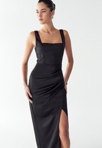 Žena v bezramínkových černých šatech se čtvercovým výstřihem a rozparkem až ke stehnům, stylizovaná s výraznými zlatými náušnicemi a náramkem.