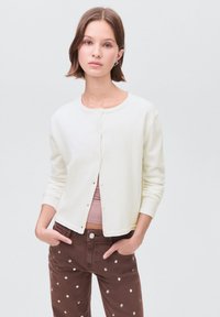 Tânără cu păr scurt, brunet, poartă un cardigan alb peste un top roz și pantaloni maroon cu aplicații argintii, stând cu mâinile în buzunare.