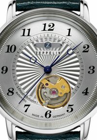 Automatische Uhr mit silbernem Sonnenschliff-Zifferblatt, blauen Zeigern, schwarzen Ziffern und sichtbar goldenem Uhrwerk. Mit einem grünen Lederarmband.