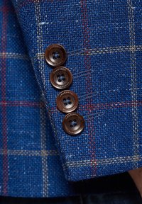 Niebieska tkanina o strukturze z wzorem w kratę z czterema brązowymi guzikami na mankiecie, prezentująca subtelny połysk i wygrawerowane detale.