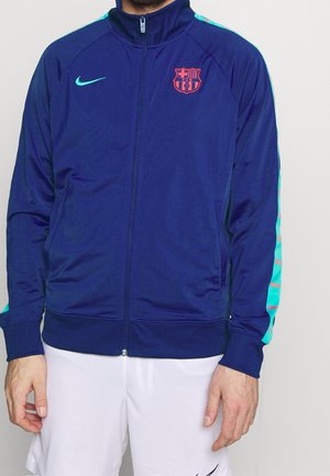 Giacca da track navy con colletto alto, chiusura con zip e tasche laterali. Presenta dettagli turchesi e il logo del Barcellona sul petto.