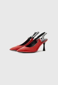 Tacones de charol rojo con punta afilada, tacón negro y correa ajustable que presenta un acento de marca en blanco y negro.