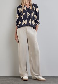 Pantaloni lunghi e beige, fluidi, abbinati a una blusa blu navy con un motivo geometrico crema, una serie di bottoni e maniche arrotolate.