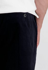 Close-up van een persoon die donkere corduroy broek draagt met een zwarte knop en een wit overhemd dat erin is gestopt, waarbij een deel van hun arm zichtbaar is.