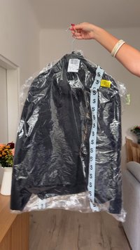 Jachetă neagră în ambalaj de plastic, cu un etichetă de culoare albastră deschisă, pe care se repetă litera 'S'. Atârnă pe un cârlig, având un autocolant de preț vizibil.