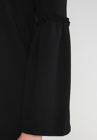 Black long-sleeve garment with ruffled detail at the cuff. Soft fabric with a smooth texture and a straight, flowing silhouette.