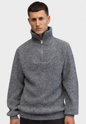 Jeune homme portant un pull en laine gris à col montant et fermeture éclair sur la moitié, debout devant un fond clair uni.