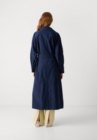 Manteau long bleu marine avec une ceinture, doté d'un large col et d'un tissu robuste, vu de dos, associé à un pantalon jaune clair.