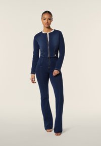 Denim overall in donkerblauw, met een cropped top met lange mouwen, een rits aan de voorkant en uitlopende pijpen. Afgewerkt met contrasterende stiksels.