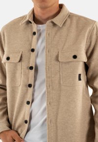 Man in een beige button-up shirtjasje over een wit T-shirt, met zwarte knopen en twee borstzakken met klep.