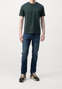 Man in een donkergroen T-shirt met korte mouwen, blauwe spijkerbroek en zwart-beige sneakers, staand tegen een effen witte achtergrond.