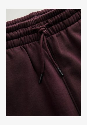 Bordo krāsas sporta bikses ar elastīgu vidukļa jostu, regulējamu auklu un teksturētu audumu. Auklas galiem melni metāla uzgaļi.