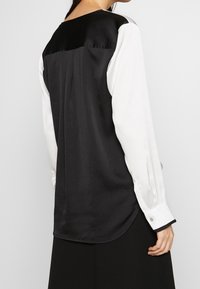 Woman wearing a black sleeveless top over a long-sleeve white shirt with buttoned cuffs, shown from the back against a plain background.