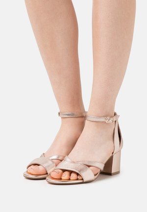 Des pieds portant des sandales à talons carrés en métal rose doré avec des brides autour des chevilles et des orteils ouverts sur un fond blanc.