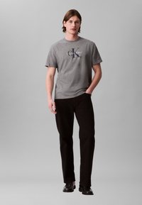 Γκρι κοντομάνικο T-shirt με κεντρικό λογότυπο "Calvin Klein" σε μπλε χρώμα. Συνδυασμένο με μαύρο παντελόνι και μαύρα παπούτσια, σε ουδέτερη στάση.