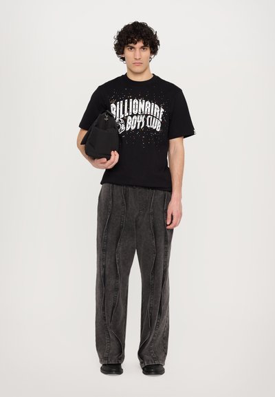 T-shirt noire avec imprimé coloré, associé à un jean baggy gris avec des coutures ondulées, et un petit sac noir tenu dans le bras gauche.